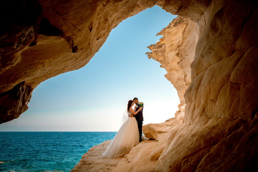 Bride and groom standing on a cliff arch overlooking the sea and blue sky in Crete – Wedspace destination wedding