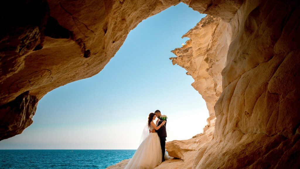 Bride and groom standing on a cliff arch overlooking the sea and blue sky in Crete – Wedspace destination wedding