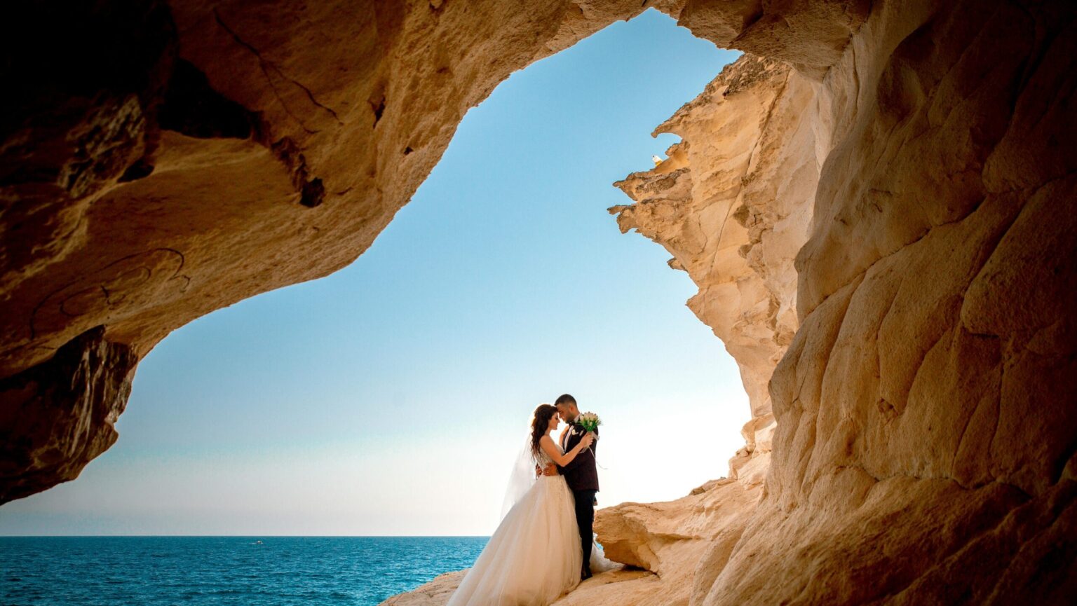 Bride and groom standing on a cliff arch overlooking the sea and blue sky in Crete – Wedspace destination wedding