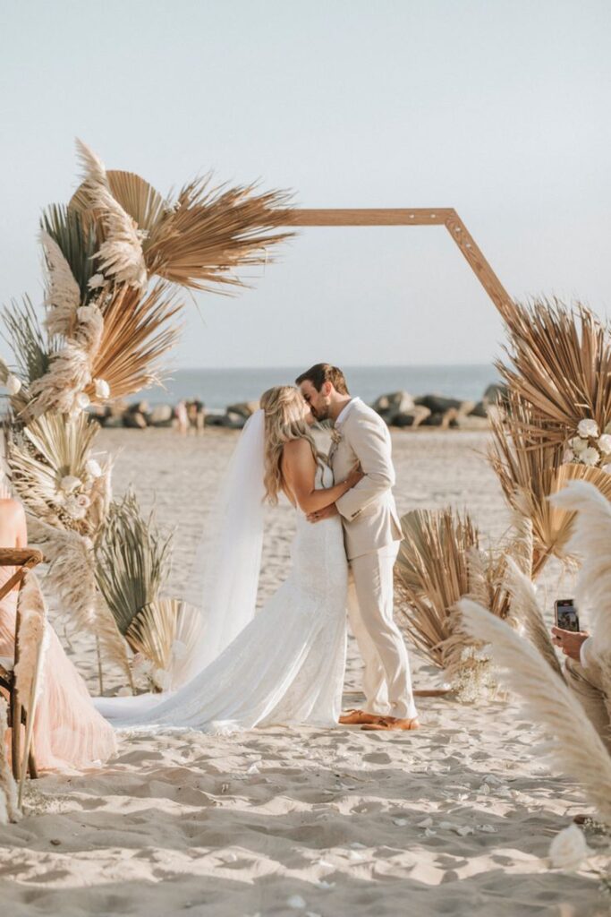 vow renewal A loving couple barefoot on the sand by a hexagonal arch with the sea in the background, sharing a kiss. Romantic vow renewal on the beach in Crete.
