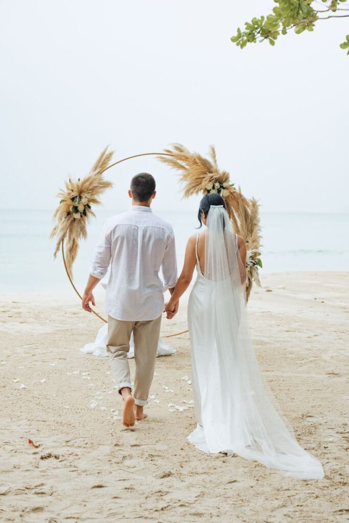 elopement A couple walking barefoot on the sand through a round decorated arch toward the sea. She is wearing a wedding dress, he is in a white shirt and pants. Romantic elopement wedding for two in Crete.