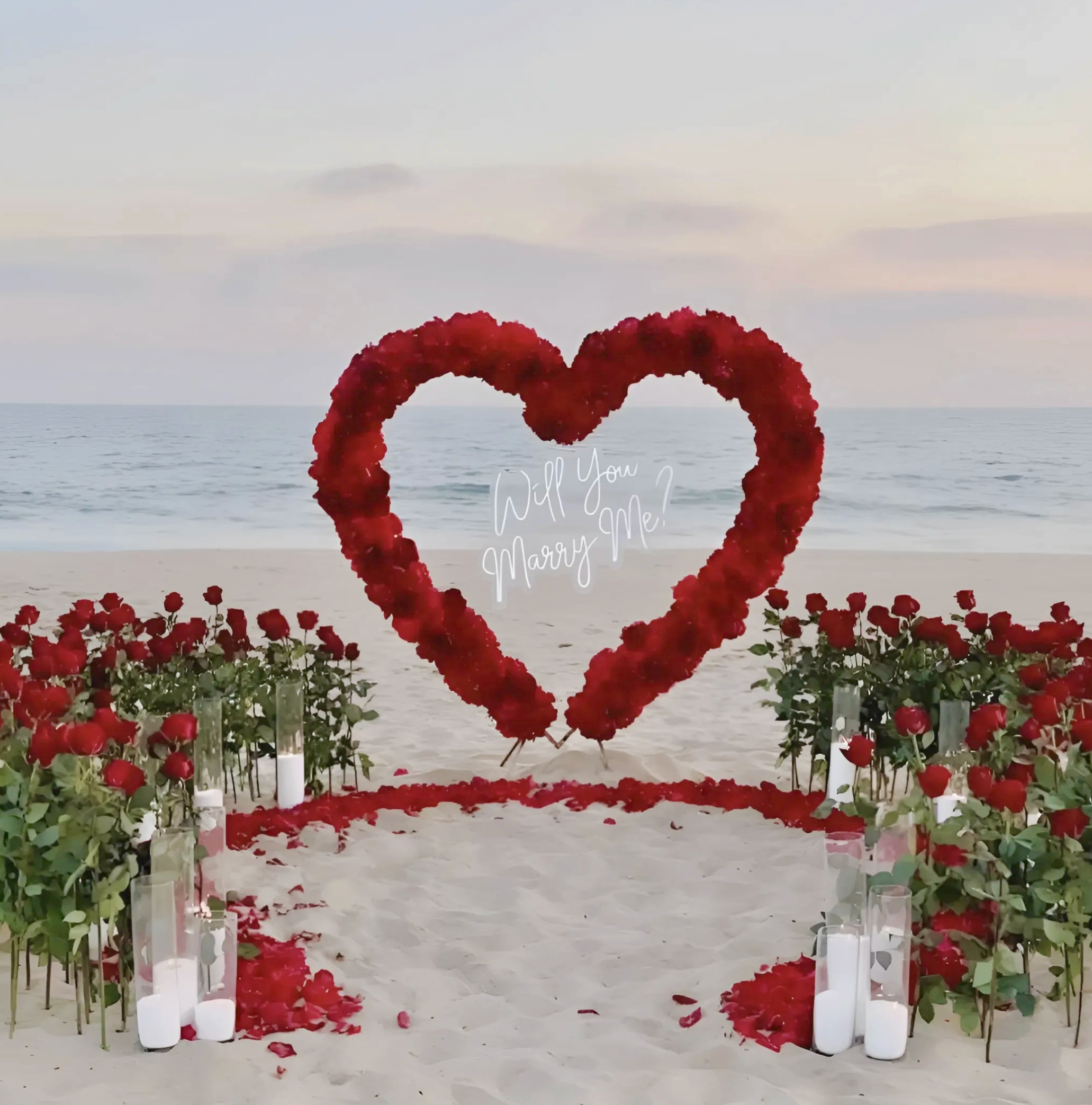 Luxury Premium marriage proposal setup on the beach in Crete - large red rose heart arch with “Will You Marry Me?” neon sign, red roses and candles on the sand at sunset