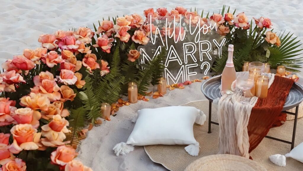Romantic beach proposal setup in Chania, Crete with roses semicircle, “Will You Marry Me?” sign, champagne and intimate picnic area on the sand