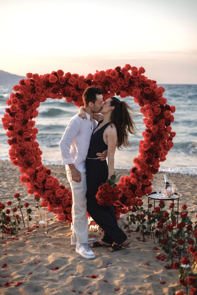 A happy couple hugging on a Cretan beach inside a large heart made of red roses, showing a Premium marriage proposal setup from Wedspace Crete
