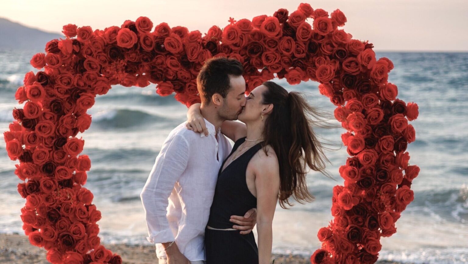 A happy couple hugging on a Cretan beach inside a large heart made of red roses, showing a Premium marriage proposal setup from Wedspace Crete