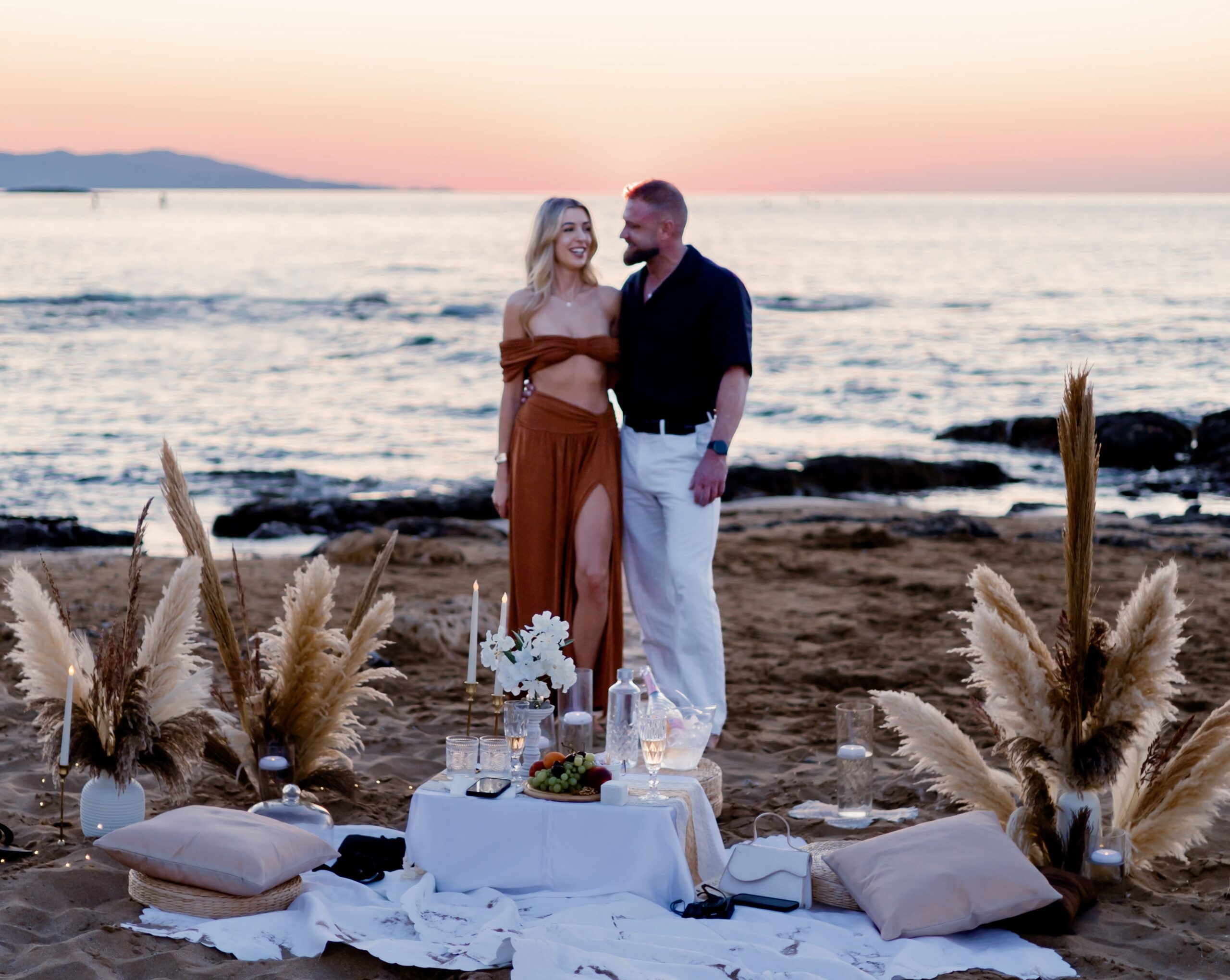 Surprise beach proposal in Crete featuring a romantic setup with pampas grass, boho decor, and sunset sea view by WedSpace Crete