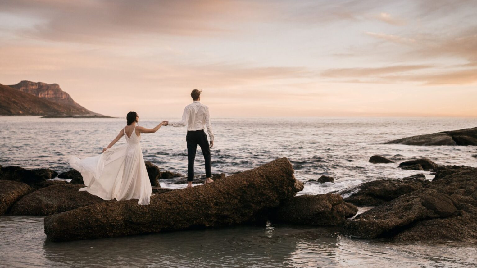 Romantic couple holding hands on coastal rocks during a sunset beach elopement in Crete, organized by Wed-Space.