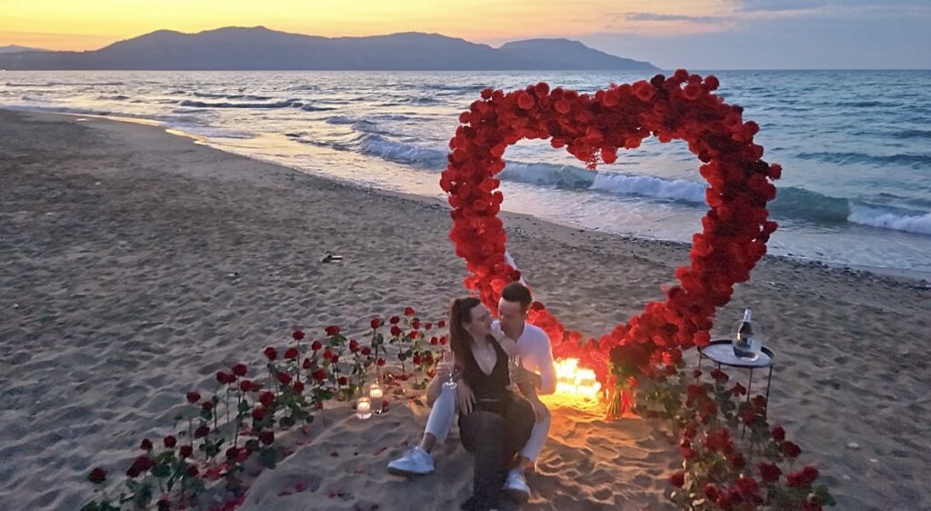 Magical golden hour view of a couple seated by a romantic rose petal path and arch on a Crete beach, organized by luxury planners WedSpace.