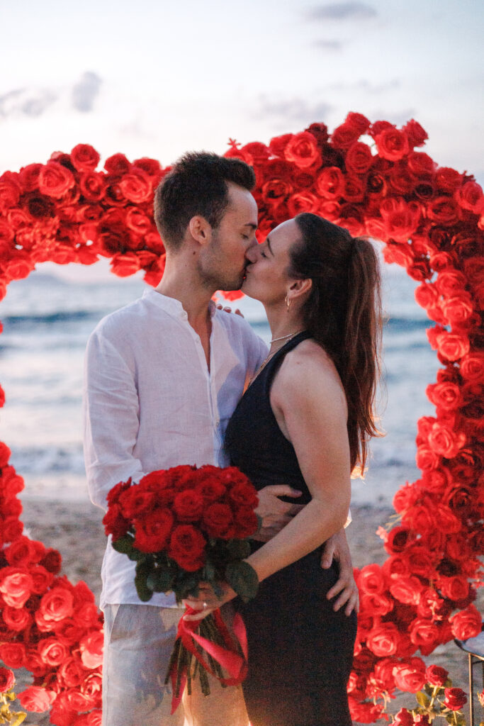 Romantic marriage proposal moment: newly engaged couple kissing inside a giant red rose heart arch on a Crete beach, planned by WedSpace.