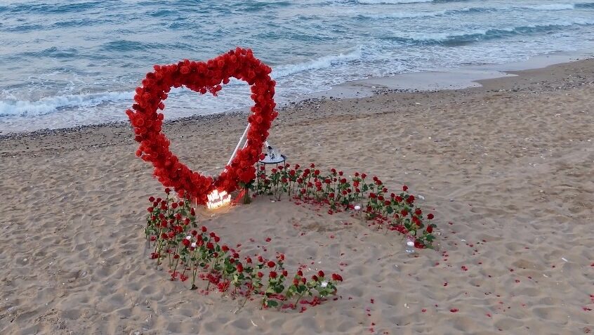 Luxury beach proposal setup in Georgioupoli, Rethymno, Crete, featuring a massive red rose heart arch and candles on the sand by WedSpace.