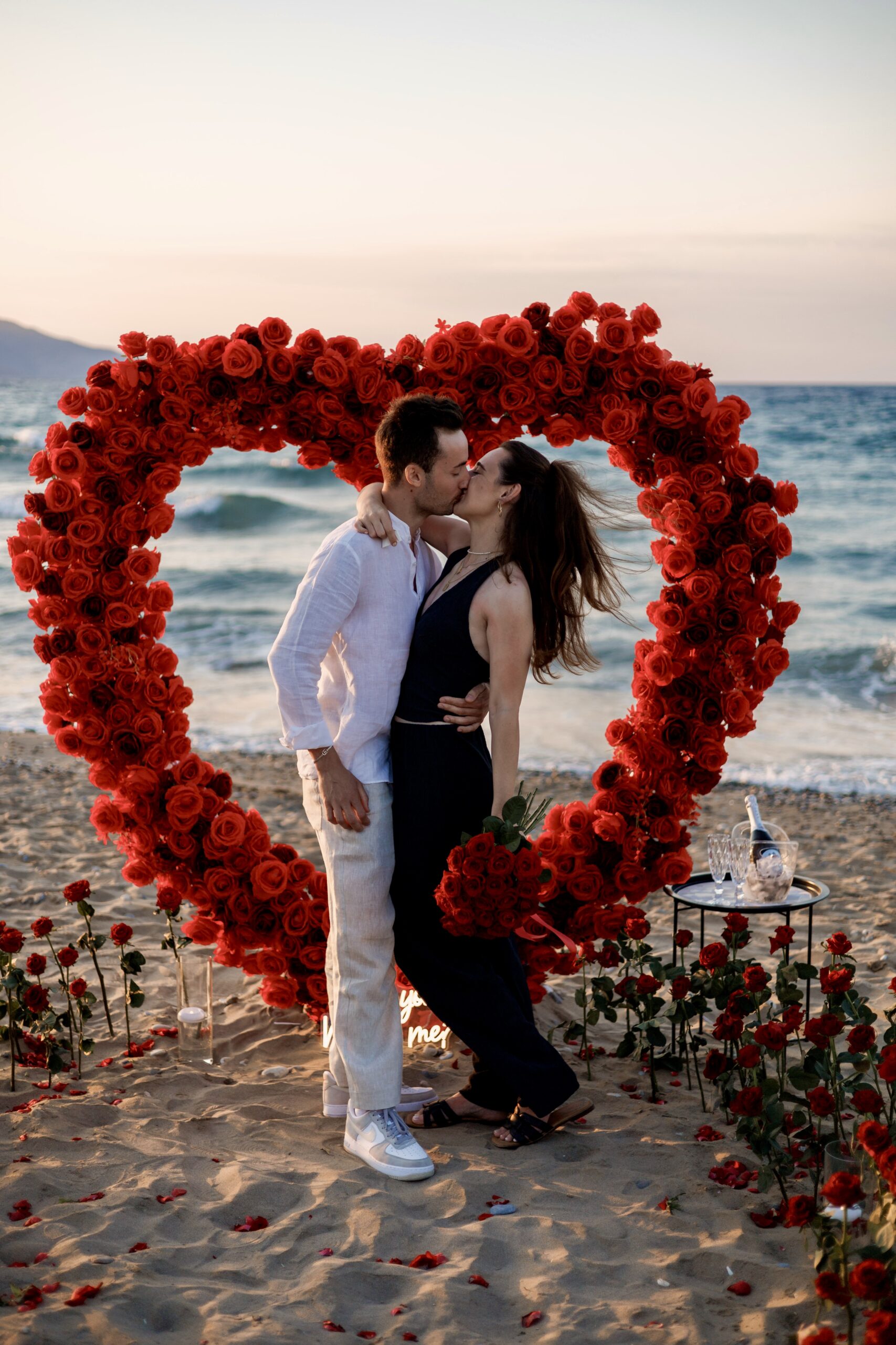 Engaged couple standing together inside a stunning floral heart arch on the shore in Crete, captured by proposal planners WedSpace.