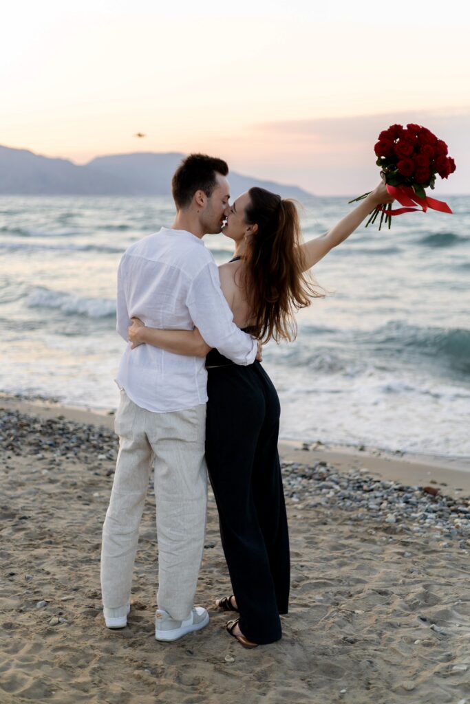 Happy couple sharing a romantic moment with a large bouquet of red roses on a Crete beach, a premium proposal by WedSpace.