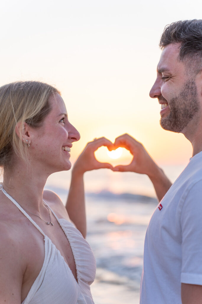 Romina and Keno making a heart shape with hands against the sunset after beach proposal in Rethymno, Crete by WedSpace Crete⁠