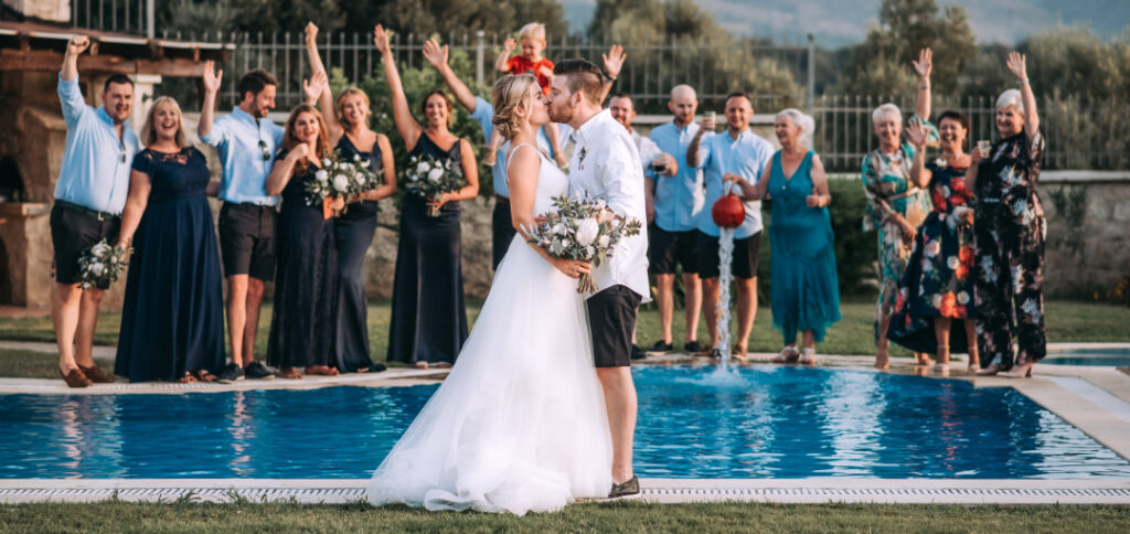⁠Bride and groom kissing by the pool with cheering guests at a small intimate wedding venue on Villa Vasilis in Crete⁠ | WedSpace