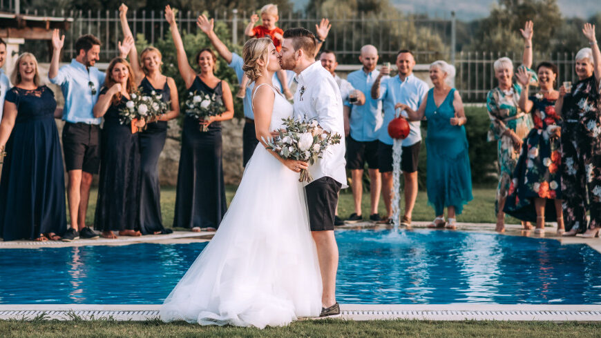 ⁠Bride and groom kissing by the pool with cheering guests at a small intimate wedding venue on Villa Vasilis in Crete⁠ | WedSpace