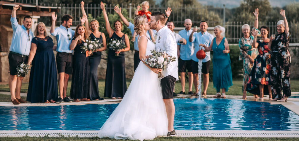 ⁠Bride and groom kissing by the pool with cheering guests at a small intimate wedding venue on Villa Vasilis in Crete⁠ | WedSpace